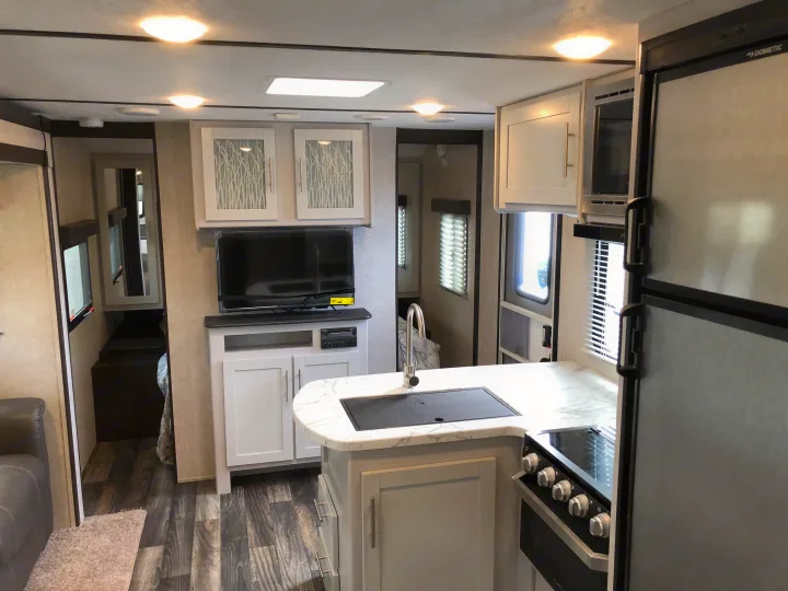 View of a modern RV rental interior showing a kitchen with sink, stove, and fridge, adjacent to a living area with a TV.. Keystone Bullet 2019