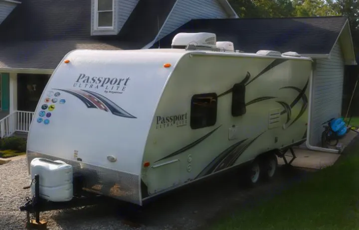A white Passport Ultra-Lite travel trailer, perfect as an RV rental, parked on gravel. Features include front propane tanks and rooftop AC units.. Keystone Passport 2014