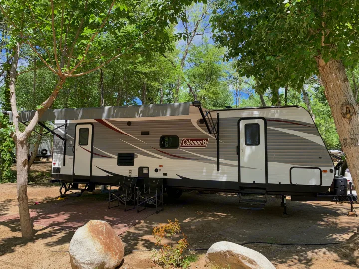 Spacious RV rental parked under shady trees at a sunny campsite with chairs, rocks, and an awning extended.. Coleman Lantern 2018