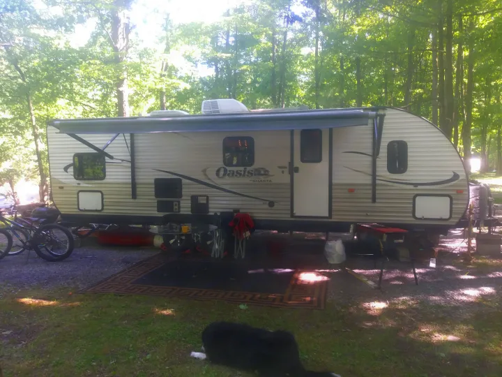 Our serene RV rental campsite. A Shasta Oasis travel trailer, with awning, bikes, and a dog relaxing, surrounded by trees.. Shasta Oasis 2013
