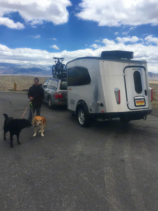 An Airstream rv rental, SUV with bikes, man, and two dogs on a scenic road trip with snowy mountains.. Airstream Base Camp 2017
