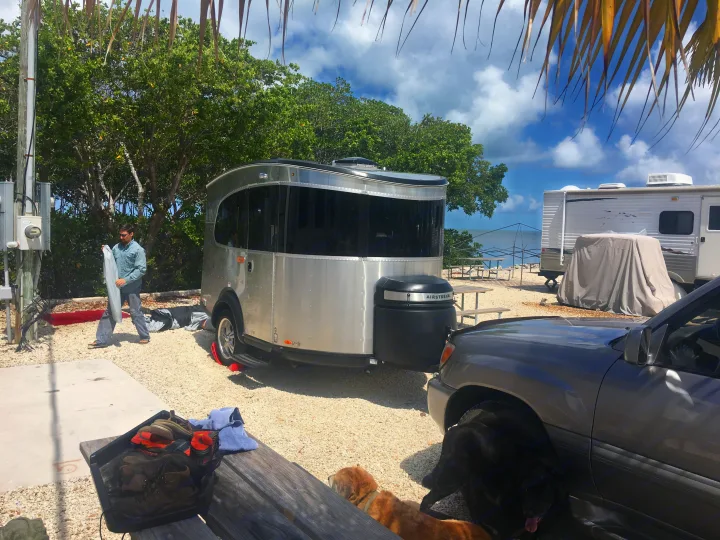 Man prepares Airstream RV rental at a sunny, gravel beachfront campground with dogs and other RVs.. Airstream Base Camp 2017