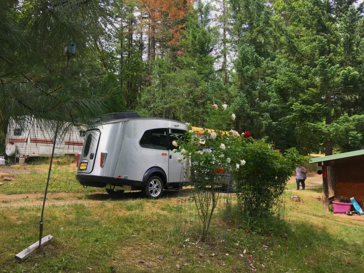 Silver compact camper and a large travel trailer parked among trees. Rose bushes and a person complete the rustic rv rental scene.. Airstream Base Camp 2017