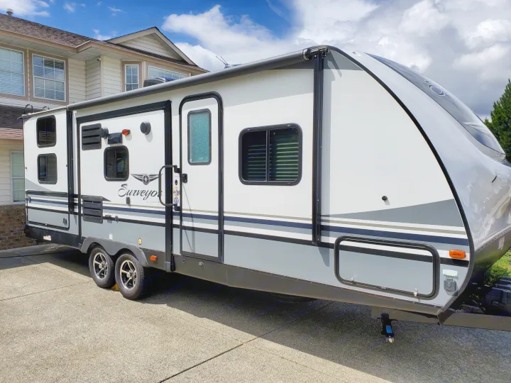 A modern white and grey travel trailer, likely an RV rental, parked on a sunny paved driveway next to a house.. Forest River Surveyor 2019