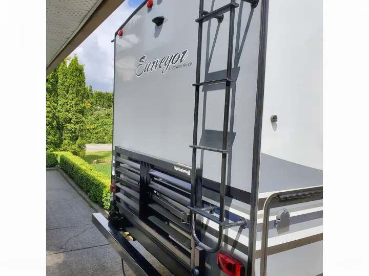 Rear of a grey Surveyor RV, highlighting its ladder and bottom storage rack. This type of vehicle is popular for rv rental.. Forest River Surveyor 2019