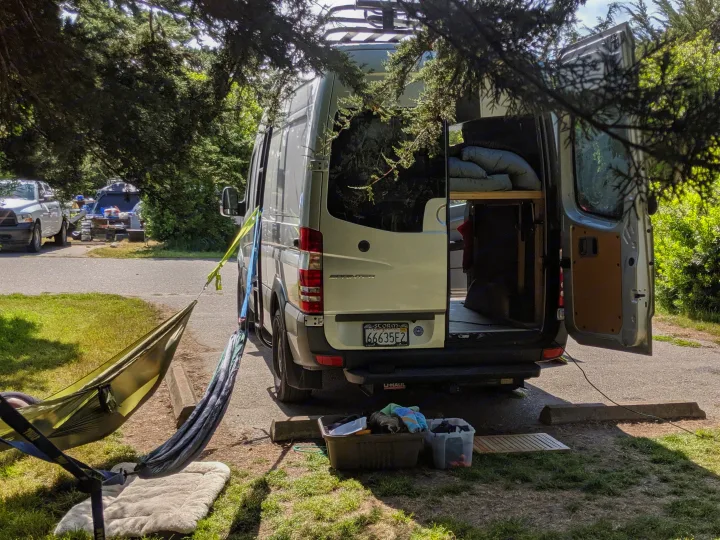 RV rental Sprinter van parked at a tree-shaded campsite. Its open rear doors show the converted interior, with a hammock outside.. Mercedes-Benz Sprinter 2016
