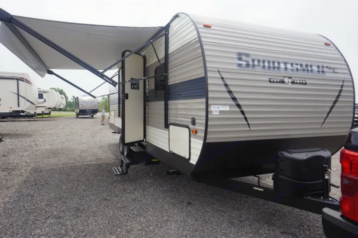 A gray Sportsmen travel trailer for rv rental, with its awning extended and door open, parked on a gravel lot.. K-Z Manufacturing sportsmen 301 2020