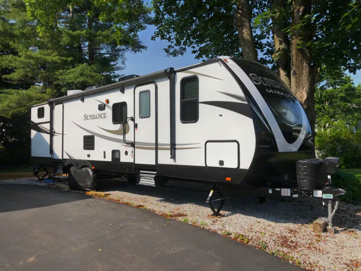 A white Sundance travel trailer, an RV rental, is parked on gravel with stabilizers down, shaded by large trees.. Heartland Sundance 2019