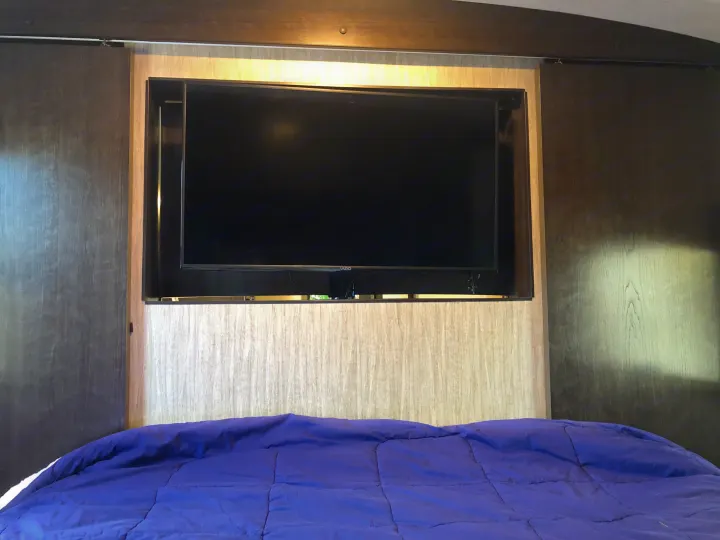 Inside this RV rental bedroom: a mounted TV above a purple bed, flanked by dark sliding doors. Ideal for cozy travels.. Heartland Sundance 2019