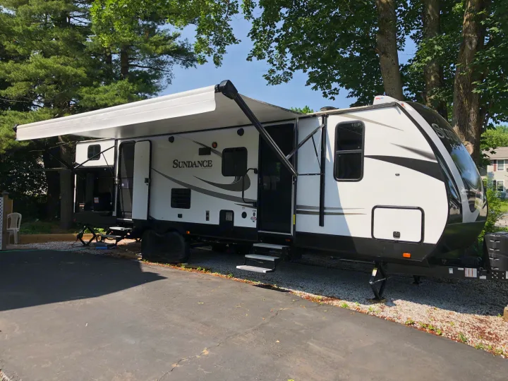 White Sundance travel trailer RV rental, awning extended, parked under trees for your outdoor adventure.. Heartland Sundance 2019