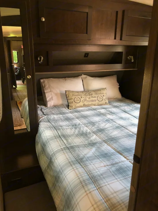 Cozy RV rental bedroom with dark wood cabinets, a plaid bed, and a mirrored closet reflecting the hallway.. Heartland Sundance 2019