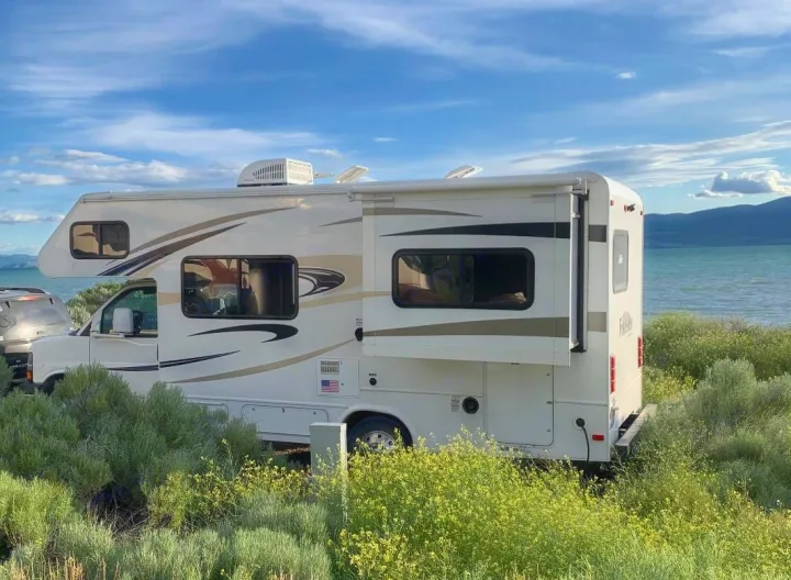 White RV rental with slide-out extended, parked by a beautiful lake with mountains. Surrounded by green and yellow bushes.. Forest River Forester 2015