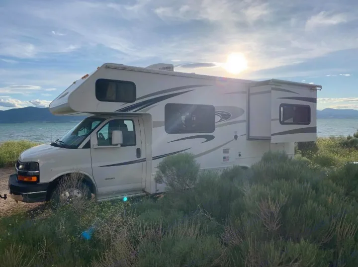 Scenic RV rental adventure! White Class C RV by a lake with mountains, under a sunny sky. Perfect for outdoor travel & camping.. Forest River Forester 2015