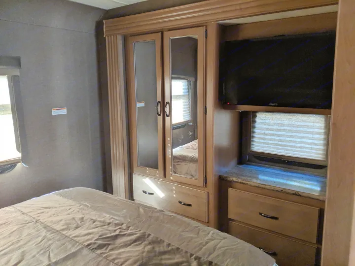 Cozy RV rental bedroom showing a bed, mirrored wardrobe, mounted TV, and windows with blinds.. Thor Motor Coach A.C.E 2019
