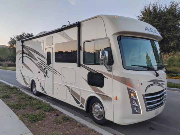 Explore with this modern beige ACE motorhome, ideal for RV rental road trips. Parked on a quiet street at dusk.. Thor Motor Coach A.C.E 2019