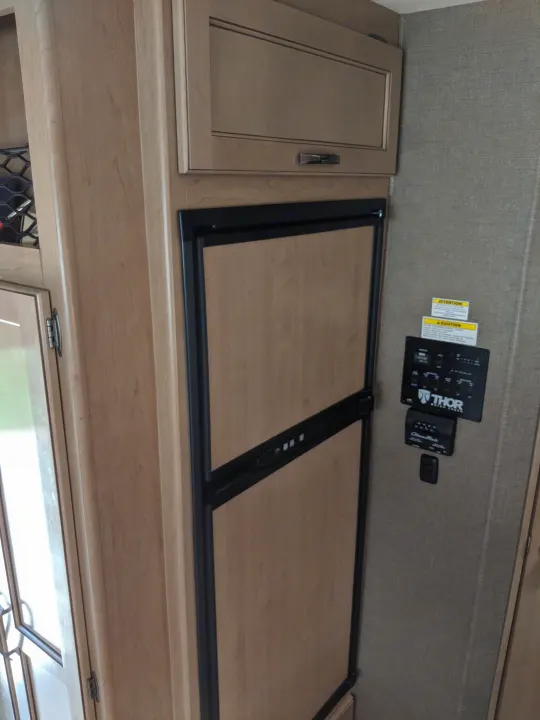 RV rental interior featuring wood-paneled refrigerator, overhead cabinets, and a control panel for monitoring systems.. Thor Motor Coach A.C.E 2019