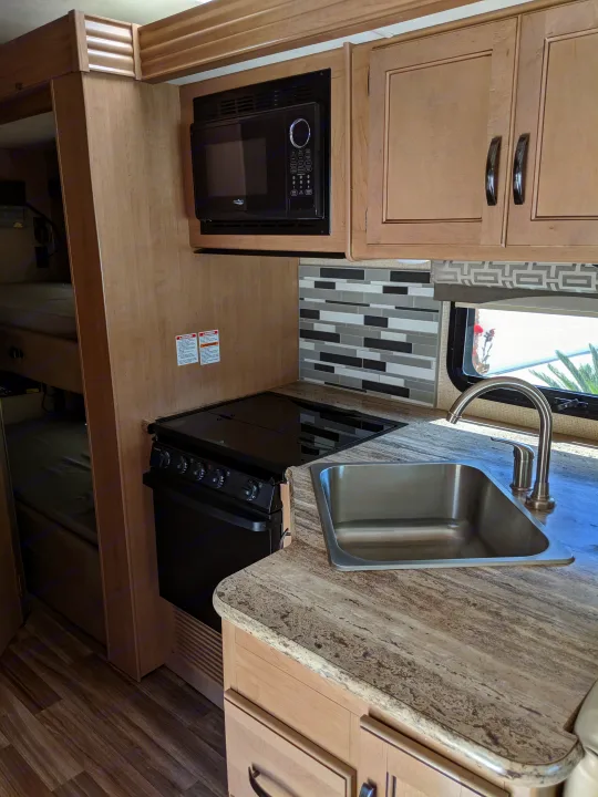 Inside an RV rental, showing the compact kitchen with microwave, stove, sink, stylish backsplash, and visible bunk beds.. Thor Motor Coach A.C.E 2019