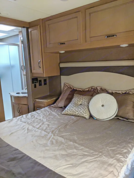 Explore this cozy RV rental's bedroom, featuring a comfortable bed, overhead storage, and a convenient en-suite bathroom with a sink.. Thor Motor Coach A.C.E 2019