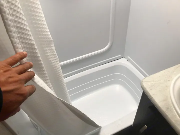 Inside an RV rental: a hand holds back a white shower curtain, revealing a compact, white shower/tub combo with integrated walls.. Keystone Outback 2019