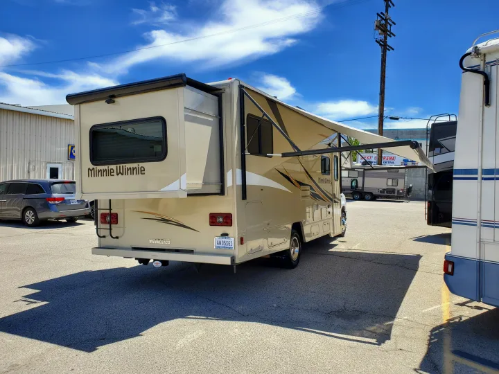 A beige Winnebago "Minnie Winnie" RV with an extended awning and slide-out, parked in a lot. Perfect for an RV rental getaway.. Winnebago Minnie Winnie 2018