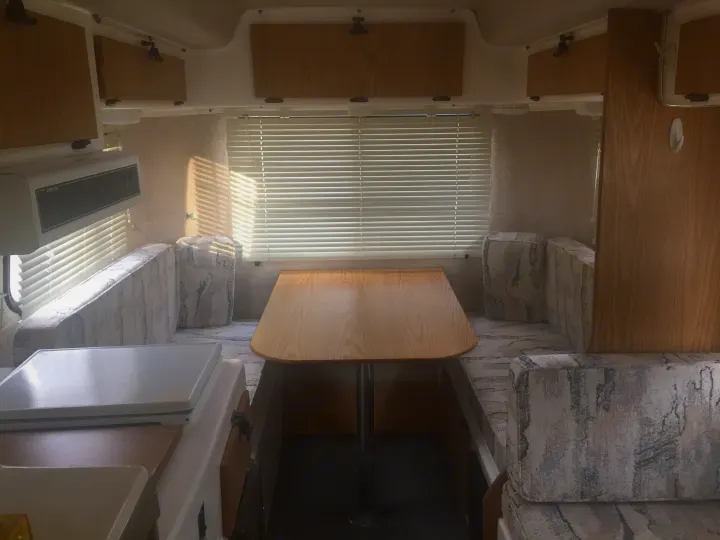 Bright RV rental interior featuring a wooden dining table, upholstered benches, overhead cabinets, and window blinds.. Casita 17' Spirit Deluxe 2003
