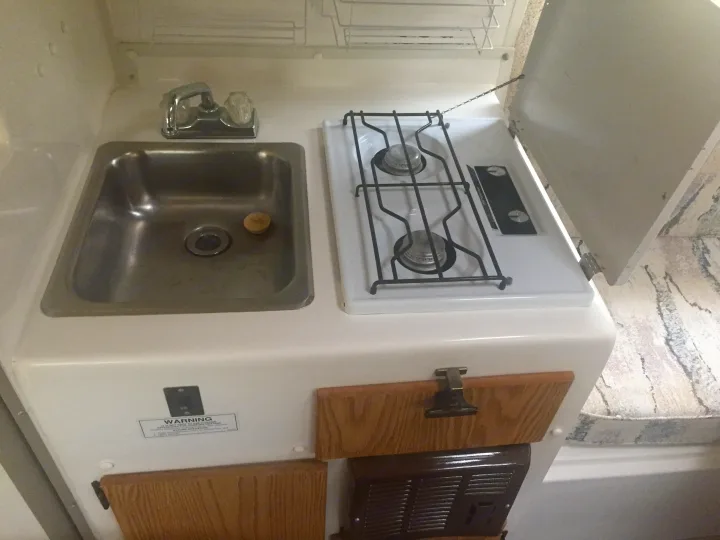 Compact RV rental kitchen with a stainless steel sink, faucet, and a two-burner gas stove with its cover open.. Casita 17' Spirit Deluxe 2003
