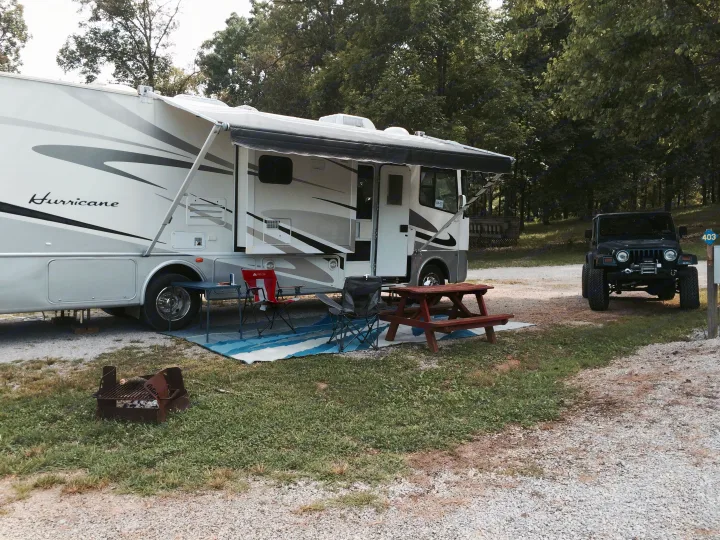 Spacious rv rental at a leafy campsite, with an extended awning, picnic setup, fire pit, and a parked Jeep.. Thor Motor Coach Hurricane 34N 2006