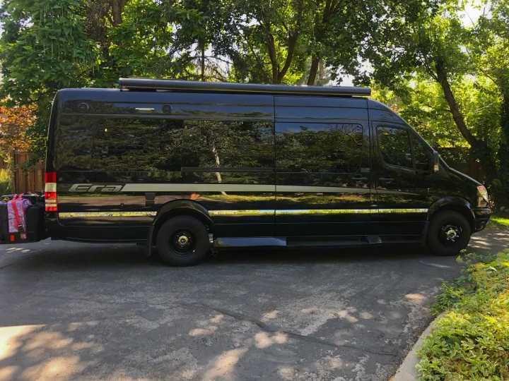 Black Winnebago Era RV rental parked on a driveway with luggage. Sunlight reflects off its side, surrounded by trees.. Winnebago Era 2014