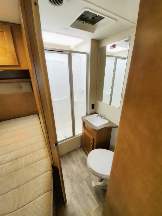 Clean RV rental interior featuring a compact bathroom with shower, toilet, and sink, adjacent to a cozy bed area.. Winnebago Minnie Winnie 2018