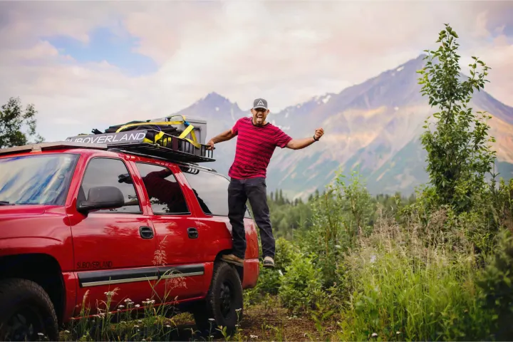 A man cheers, standing on a red "SUBOVERLAND" SUV with mountain backdrop. Experience adventure like this with an RV rental.. Chevrolet Suburban Overland 2001