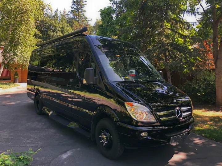 Black Mercedes-Benz Sprinter RV rental camper van parked on a sunny driveway amidst green trees.. Winnebago Era 2014