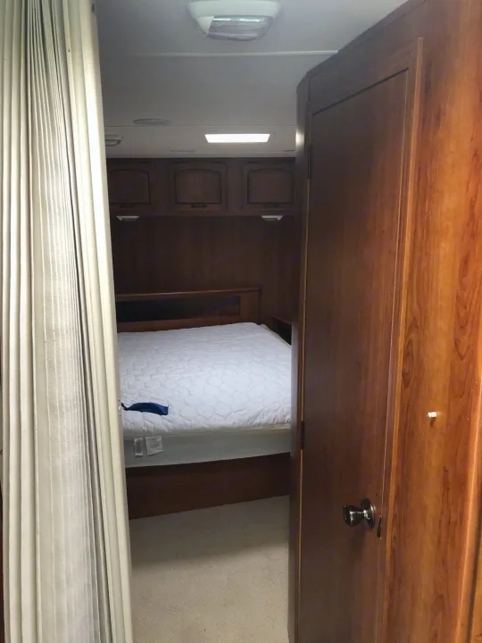 View of an RV rental bedroom featuring a white quilted bed, wooden overhead cabinets, and a privacy curtain. A wooden door is on the right.. Ford Coachman 2009