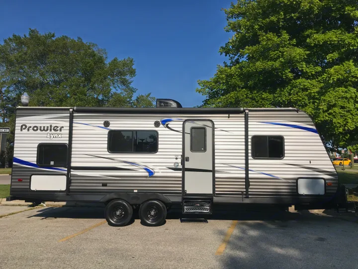 Prowler Lynx travel trailer RV rental, white/gray with blue accents, parked on asphalt under trees & blue sky.. Heartland Prowler Lynx 2019