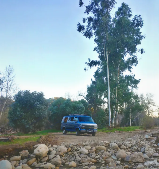 Blue RV rental parked by a rocky riverbed & tall trees in a serene natural setting.. Chevrolet Other 1989