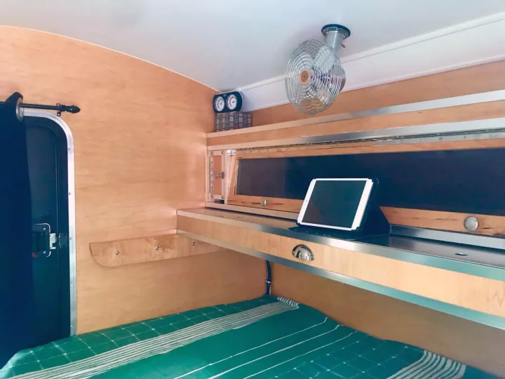 Step inside this cozy RV rental with light wood interior, green bedding, and fan. Tablet on shelf, compact camper living.. The Teardrop Trailer. N/A 2019