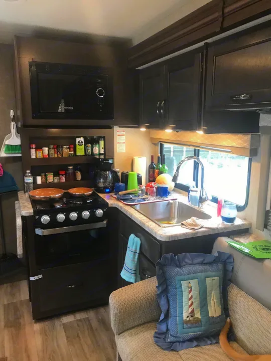 Inside an RV rental, a functional kitchen with stove, microwave, spice rack, and sink with a scenic water view. Cozy camper life.. Thor Freedom Elite 2020