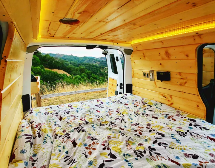 Step into this cozy RV rental's wooden interior, offering a comfy bed and scenic nature views from the open back.. Nissan NV200 2019