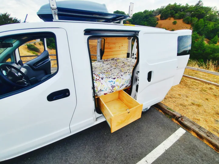 Custom white camper van with a cozy bed, pull-out storage, and roof box. Ready for your next RV rental adventure!. Nissan NV200 2019