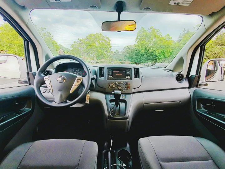 Inside an rv rental van. The driver's view reveals the dashboard, steering wheel, and the road ahead through the windshield.. Nissan NV200 2019