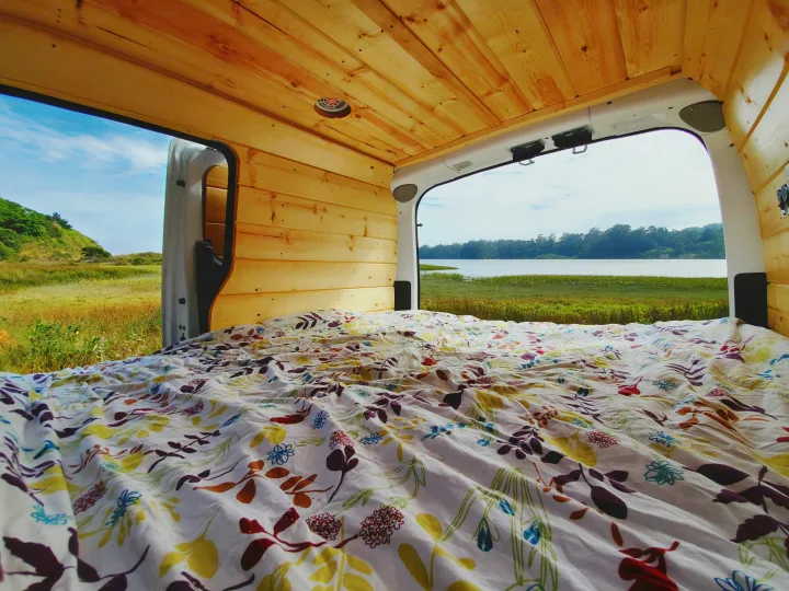 Inside a custom rv rental van, a comfy bed faces open doors with a beautiful view of a lake and grassy hills.. Nissan NV200 2019