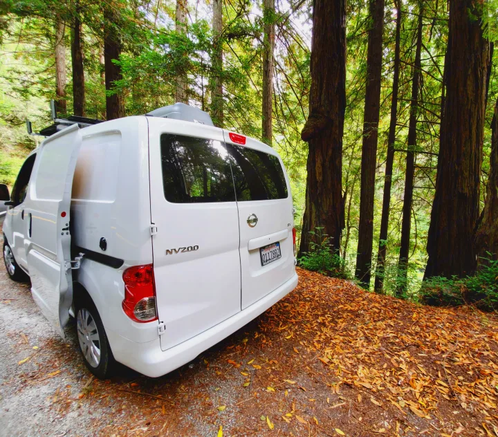 A white Nissan NV200 RV rental, with open rear doors, is parked on a leaf-strewn forest path amidst tall trees.. Nissan NV200 2019