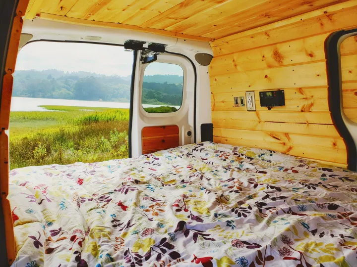 RV rental view: Cozy van interior with wooden walls, floral bed, and stunning lake landscape out the back doors.. Nissan NV200 2019