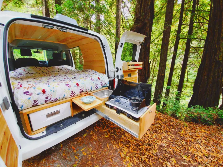 Inside a white camper van RV rental: a floral bed & pull-out kitchen with stove/sink, parked in a lush forest.. Nissan NV200 2019