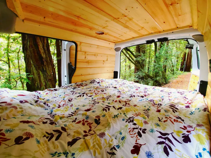 Cozy bed inside a wooden rv rental camper van, with open doors showcasing a majestic redwood forest.. Nissan NV200 2019