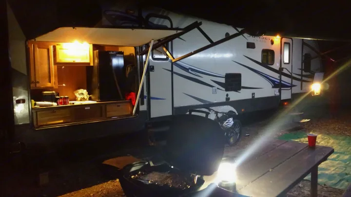 Nighttime camping scene with an RV rental, featuring a brightly lit outdoor kitchen, a grill, and a picnic table setup.. Crossroads Sunset Trail Reserve 2014