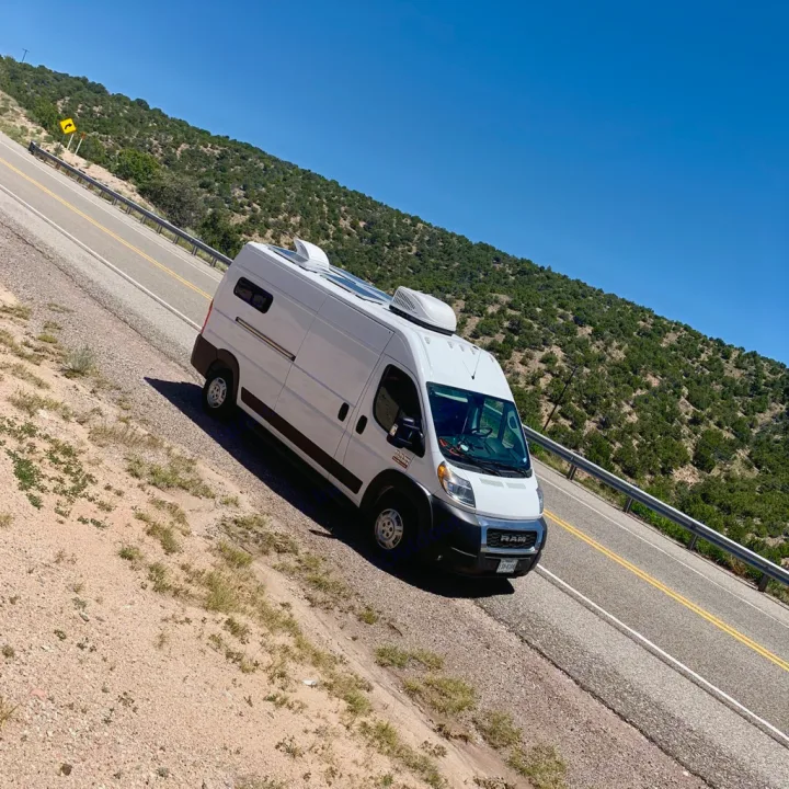 Bright white camper van RV rental equipped with solar panels on a scenic hillside road under a clear blue sky.. Dodge 2500 Promaster 2019