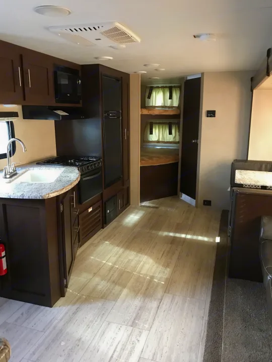 Modern RV rental interior with a functional kitchen (sink, stove, fridge), built-in bunk beds, and a living area. Light flooring and dark cabinets.. K-Z Manufacturing Sportsmen 2018