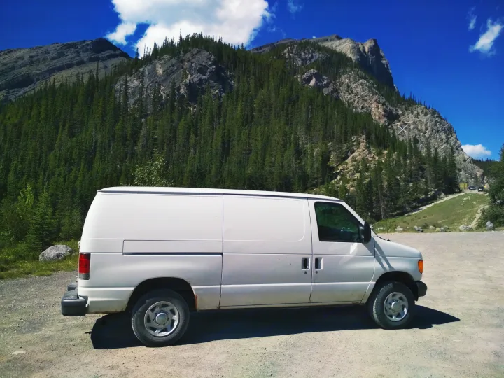 A white **RV rental** van rests on a dirt lot, framed by majestic mountains, lush evergreen forests, and a bright blue sky.. Ford Econoline 2006