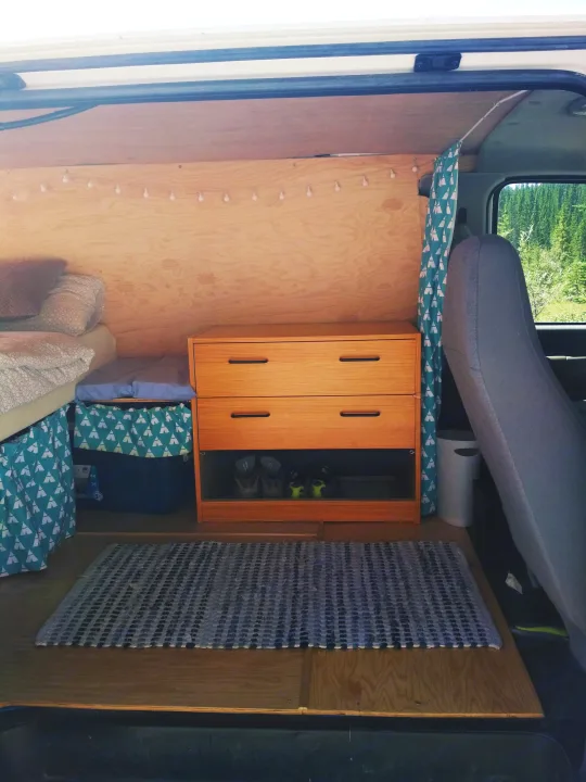 Inside a cozy RV rental van: custom plywood interior with bed, dresser, storage, patterned curtain, and forest views.. Ford Econoline 2006