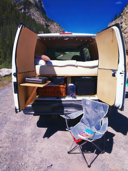 Explore nature in an rv rental! This van conversion shows a comfy bed, camp stove, & chair against scenic mountains.. Ford Econoline 2006